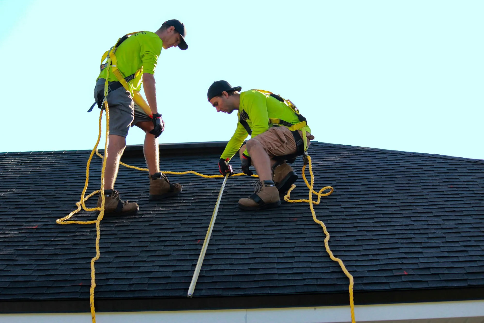 Two roofers in harnesses and hi-vis shirts measuring along a dark shingle roof
